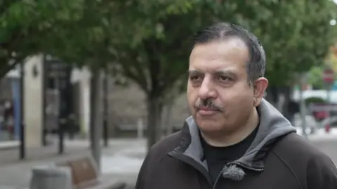 An image of Mohammed Iqbal MBE, he's wearing dark clothing and is standing in the street outside Nelson Town Hall