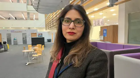 A woman wearing glasses and a blazer. She is stood in council offices.