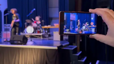 Andrew Turner/BBC A mobile phone seen on the right of the image, depicts the act on stage, seen in the left of the image. A hand is holding the phone, and two boys are playing music on the stage at St George's Theatre in Great Yarmouth.