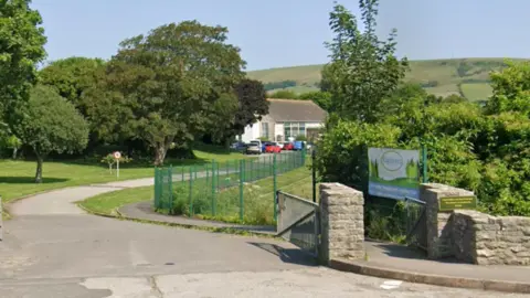 Google Google Street View of St Mark's Primary School set against the backdrop of the Purbeck Hills. A long sweeping drive is bordered by grass and mature trees. At the end is a white single-storey with about half a dozen cars parked outside.
