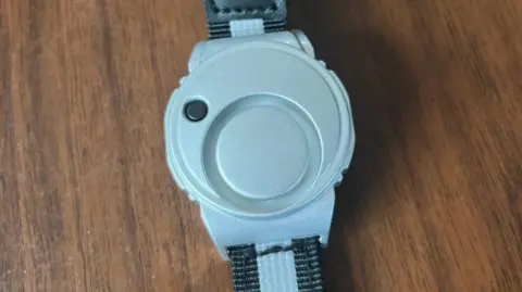 A running wristband which has a black strap with a silver plastic round watch type attached. On this is a small black button you press to sound the alarm