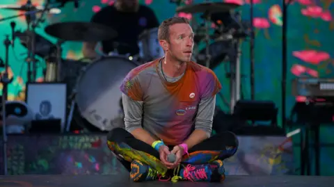 PA Media/Peter Byrne Chris Martin of Coldplay, he sits on the stage holding a microphone. He is wearing an outfit covered in ultra-violet paint splatters.