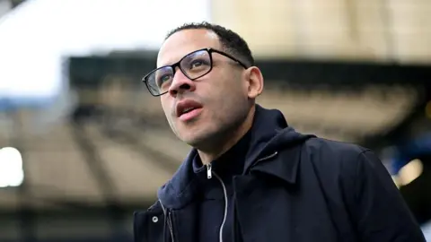 Close-up of Liam Rosenior. He has short black hair and is wearing black-rimmed glasses and a navy coat.
