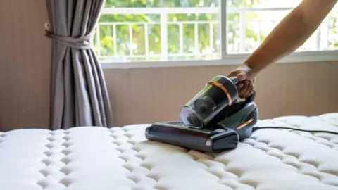 Getty Images A grey mini-vacuum device slides across a white mattress on a bed. There are grey curtains and a white window frame in the background.