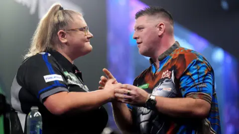 PA Darts player Beau Greaves shaking the and of another player and smilinh. She ha blonde hair in a ponytail and glasses 