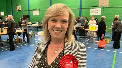 Labour Councillor Sharon Harvey, the leader, is stood at the election count wearing a red Labour rosette. Vote counting staff are standing at a number of tables in a sports hall that has a blue floor and green walls.