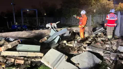 Firefighters surrounded by rubble from a collapsed building. There are two people in the picture who are wearing hi-vis uniform and helmets.