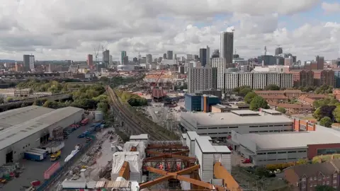 An aerial view showing the construction of a section of railway viaduct, with struts holding it up and workers in orange fluorescent uniforms. Tall city buildings can be seen in the distance on a cloudy day. 