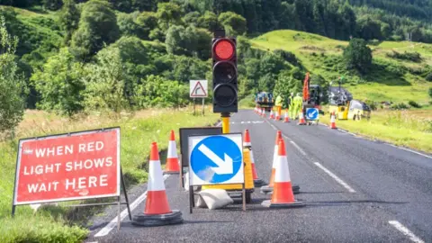A traffic light, on red, traffic cones and signs sit on a two-lane country road on a sunny day. Maintenance workers wearing high-visibility gear, as well as construction vehicles, can be seen in the distance. The road side is lined by green grass, with trees and bushes on the hills in the background. 