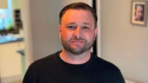 Dominic Slater Scott who has short hair and a trimmed beard, wears a dark crew-neck sweatshirt. The blurred background is shows the kitchen area of a private home.
