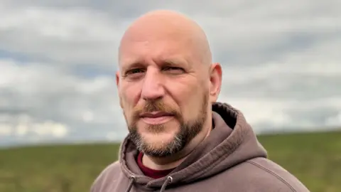 A bald headed man with ginger, dark brown and grey facial hair looks at the camera as he stands in the mostly green peatland. He is wearing a brown hoodie which has white writing in the corner.