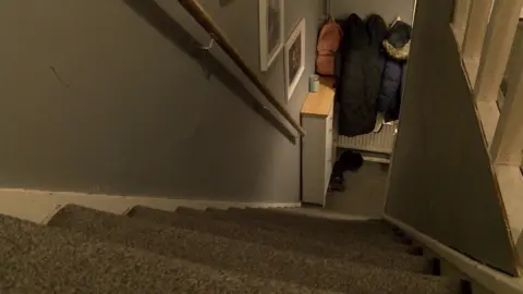 View from the top of a narrow staircase in Ayla's council house. The carpet is brown and the walls grey. Coats hang on pegs at the bottom of the stairs.
