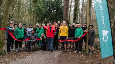 Forestry England A group of about 20 people - men and women, with one man in a wheelchair and also a black dog - standing smiling to camera in woodland. A man in the middle has just cut a red ribbon festooned between two trees. There is a turquoise upright banner saying 'Forestry England' in the foreground on the right 