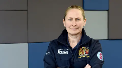 Aileen O'Connor Aileen O'Connor stood with her arms crossed. She has brown hair that is tied back, and is wearing a coat with the name of the police federation and its logo embroidered in. The wall behind her is blue and grey.