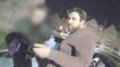 PSNI A man in a brown top looks towards the left of the screen. He has dark hair and a beard. There are houses in the background. A police officer's face is blurred.