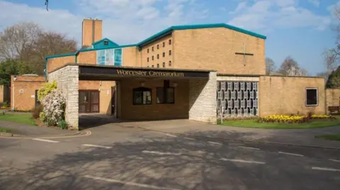 The exterior of Worcester crematorium, a brick building by the side of a road, next to a grassy patch with flowerbeds. The building has a chimney and a stained glass window is visible. A sign above the entrance reads "Worcester Crematorium".
