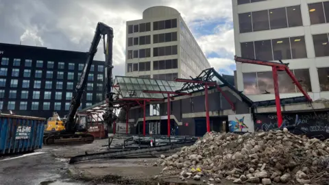 BBC Debris and rocks next to a digger in front of office buildings.