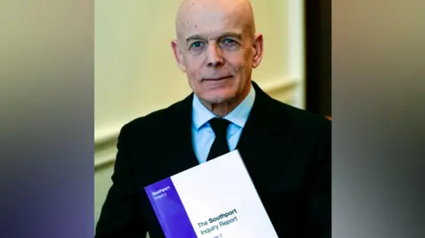 PA Media Sir Adrian Fulford, who has a bald head, is clean-shaven and wears a dark black suit, looks into the camera with a determined expression. He is holding a thick, bound copy of the Southport Inquiry Report, which has a purple and white front cover.