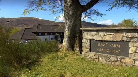 A nursing home which is obscured by a large tree in the centre of the shot and a brick wall with a plaque which says "Home Farm nursing home"