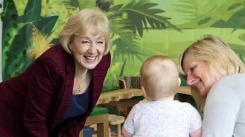 A person wearing a red and black checkered jacket is leaning forward and engaging with a young child who is dressed in a light-colored floral outfit. Another adult is seated close to the child, and the background features a vibrant jungle-themed wall with green leaves and plants. Wooden play furniture is visible in the foreground, suggesting the setting is a play area or early years environment.