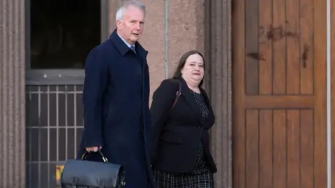 PA Media Esther Baker walking into court, she wears a black patterned dress under a black blazer. She has long brown hair. She walks alongside a man who has short grey hair. He wears a long blue coat and carries a black bag.
