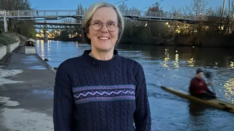 Gavin Kermack A woman with mid-length greying hair and a navy blue sweatshirt and round glasses stands on a river bank. In the background there is a footbridge and someone is paddling a kayak along the water.