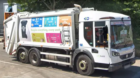 A SUEZ waste collection lorry. It is white with images on the side about recycling.