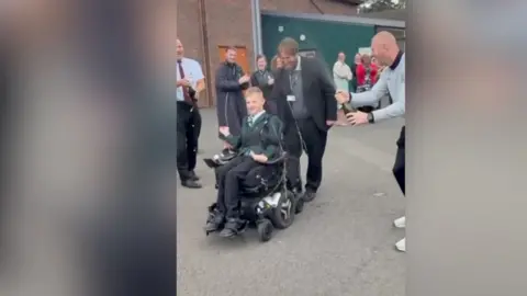 A teenager in a wheelchair, in a playground. Behind him are numerous teachers and friends.