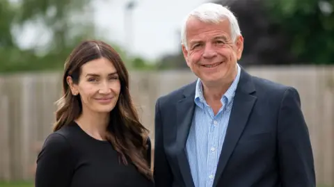 James Brew Photography Lady Peta Cavendish stands next to Chris Hall hospice chairman. They both smile. She has long, dark hair and wears a black top. Mr Hall wears a navy suit jacket and blue and white striped shirt, which is open at the collar.