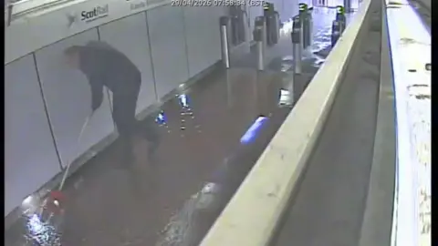 CCTV footage showing flooding in a corridor at a train station. A worker is standing amidst the water, with some sort of mop trying to contain the water.