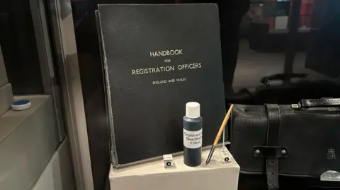 A wooden fountain pen with a black nib, a bottle of dark ink with a label reading registrars blue/black ink, a large black notebook with the words "handbook for registration officers England and Wales" in gold letters and a black leather case with "E&R" embossed on it in a display case.
