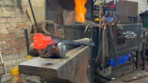 In the foreground there's a fire poker with a ram's head decoration resting on top of an anvil - the head is red-hot, it's just been in the forge. In the background you can see the forge with bright orange flames leaping up from the coals; there's tools hanging from the side, like pliers, hammers, and clamps. 