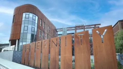 Belfast Trust The maternity hospital at the Royal Victoria Hospital. ou can see a multi-story building with copper coloured cladding and large windows. In the foreground there is grey pavement and a copper coloured sign spelling out the word maternity. 