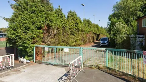 Google Locked green entrance gates to the site, with a row of conifers along the path and a van parked on the drive 
