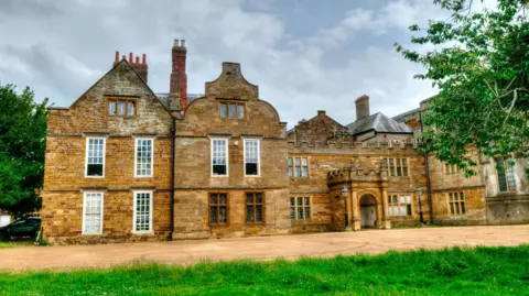 A brown, old building with grey skies and green grass in front of it.