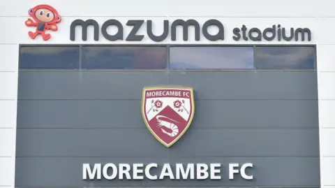 A view of the outside of Morecambe's Mazuma Stadium with the badge and 'MORECAMBE FC' emblazoned on it