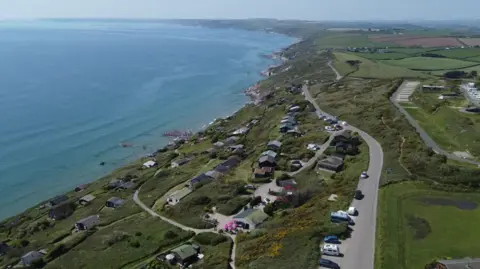 An aerial view of a coastal road. There are cars are houses on land and the blue sea on the left hand side, and open green and brown fields to the right.