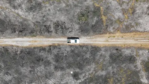 A white pick-up truck drives along a gravel track surrounded by blackened heath following the severe fire 12 months ago.