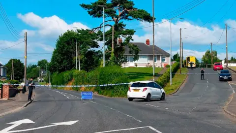 The image has been taken on the side of a road. There is a T junction leading to a bungalow on top of a small hill. A police cordon and blue sign are preventing any traffic from travelling along the main road. A small white car is parked at the bottom of the hill. An ambulance and a number of other cars are outside the house. Two police officers are standing on the road. 