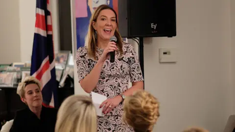 Dame Antonia Romeo has long brown hair and is wearing a floral patterned dress in pale pink and blue. She is speaking into a microphone