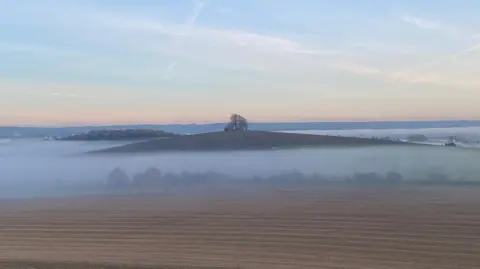 Tim A landscape view of a mist filled valley and clear skies. Rising above the mist is a barrow hill at Brightwell cum Sotwell. There is a solitary tree standing at it's peak.