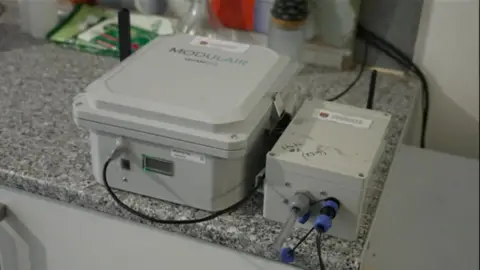 Two gray boxes on the kitchen side. One is more greedy and reads "Modular"He has a black wire that comes from him, which attaches to a wall behind her. Next to it is a smaller gray box that has a transparent tube, protruding on the side and black wires. They are machines that measure the levels of contamination