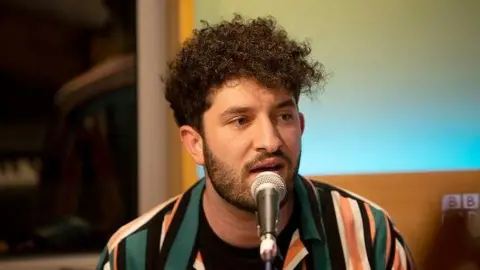 Billy Lockett with medium length dark curly hair and a short beard. He is singing into a microphone against a blue and brown background. He is wearing a jacket with green, orange and black stripes.