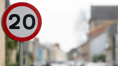 A circular white sign with a red border and the number 20 in the middle, in black lettering with a blurred street scene in the background