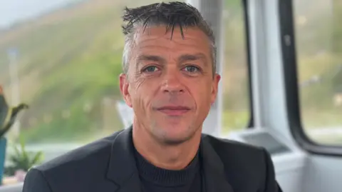 Lorne Castle smiles at the camera. He has gelled, grey hair which is short at the sides. He wears a dark jacket over a black, collarless top.