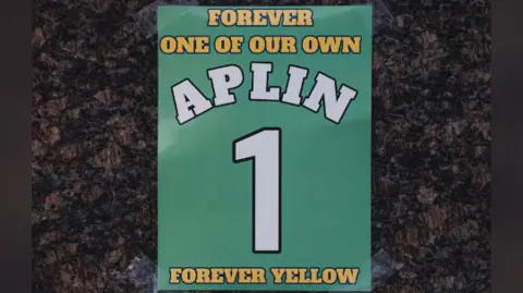 Oxford United A green poster with number 1 and the words "Forever one of our own. Forever Yellow. Alpin 1". It is taped against a marble statue base.