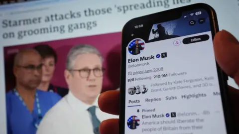 A mobile phone showing the official account of Elon Musk on social media app X (formerly Twitter), displaying a pinned post - a poll asking if 'America should liberate the people of Britain from their tyrannical government'. This held up against a computer screen displaying a BBC story headlined 'Starmer attacks those 'spreading lies' on grooming gangs, with an out-of-focus image of Keir Starmer. 