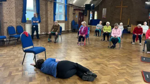 A group of people sat in a church hall on chairs holding exercise bands