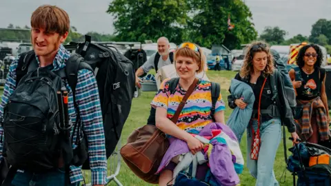 Nathan Roach/Shindig Festival People walking through a field with backpacks, bags and camping gear.