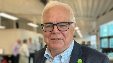 Steve Berry, an older man with short, white hair, smiles at the camera. He is wearing black-framed square glasses, a grey suit jacket and a blue and white checkered shirt. He is also wearing a green pin. He is standing in a large room with people in the background, but they are blurred.